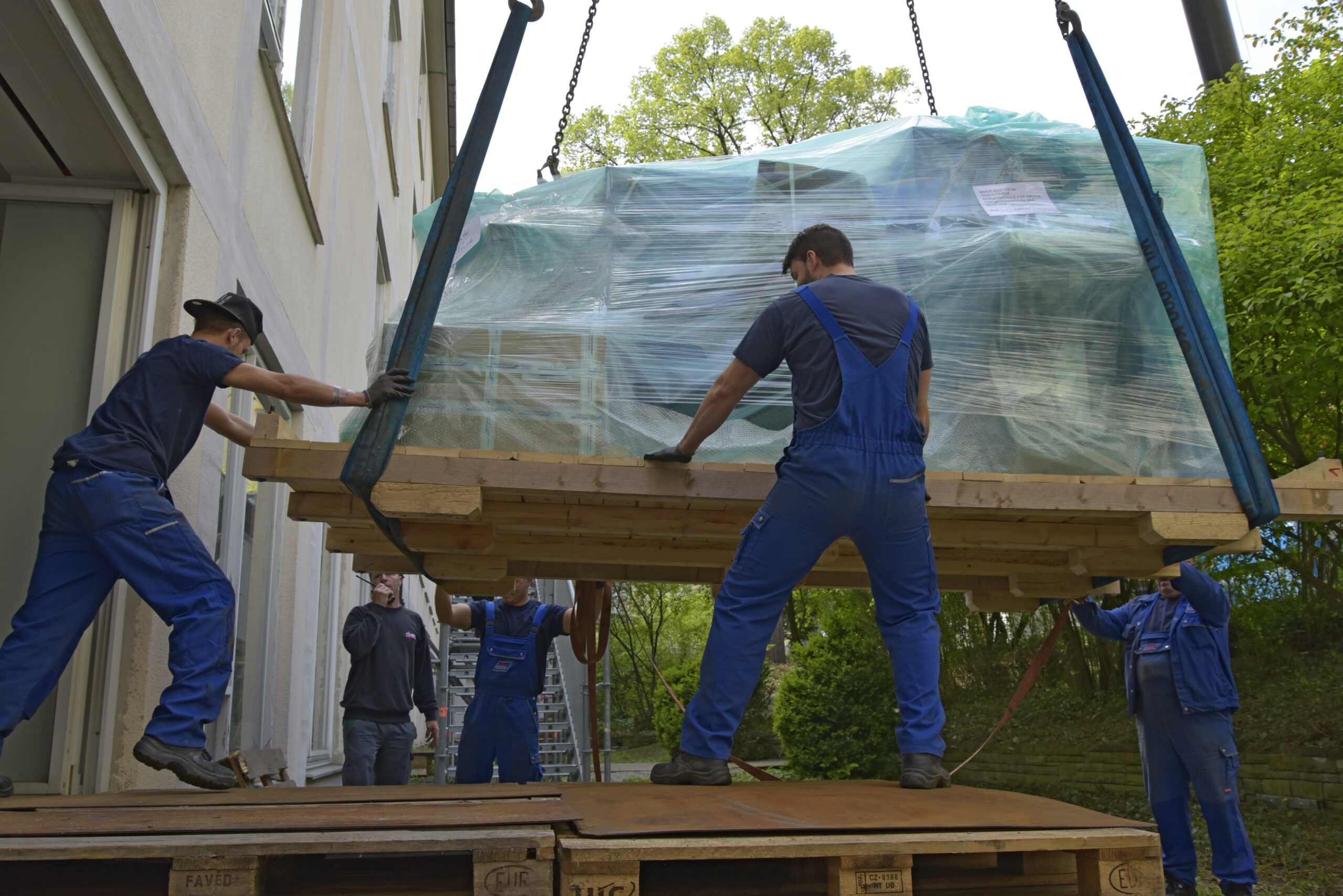 Vier Arbeiter in blauer Arbeitskleidung sichern eine große, verpackte Ladung, die mit einem Kran gehoben wird.