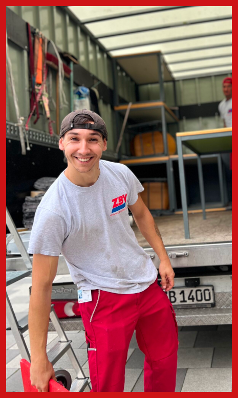 Junger Mann in roter Hose und weißem T-Shirt lächelt und steht vor einem offenen Transporter.