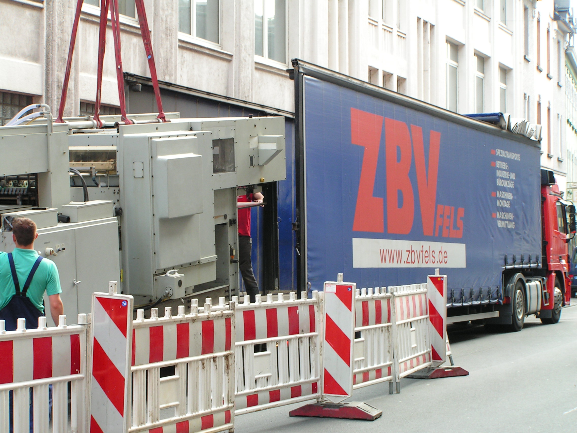 LKW mit ZBV-Logo lädt großen Transformator auf einer Straße, abgesperrt mit Bauzäunen.
