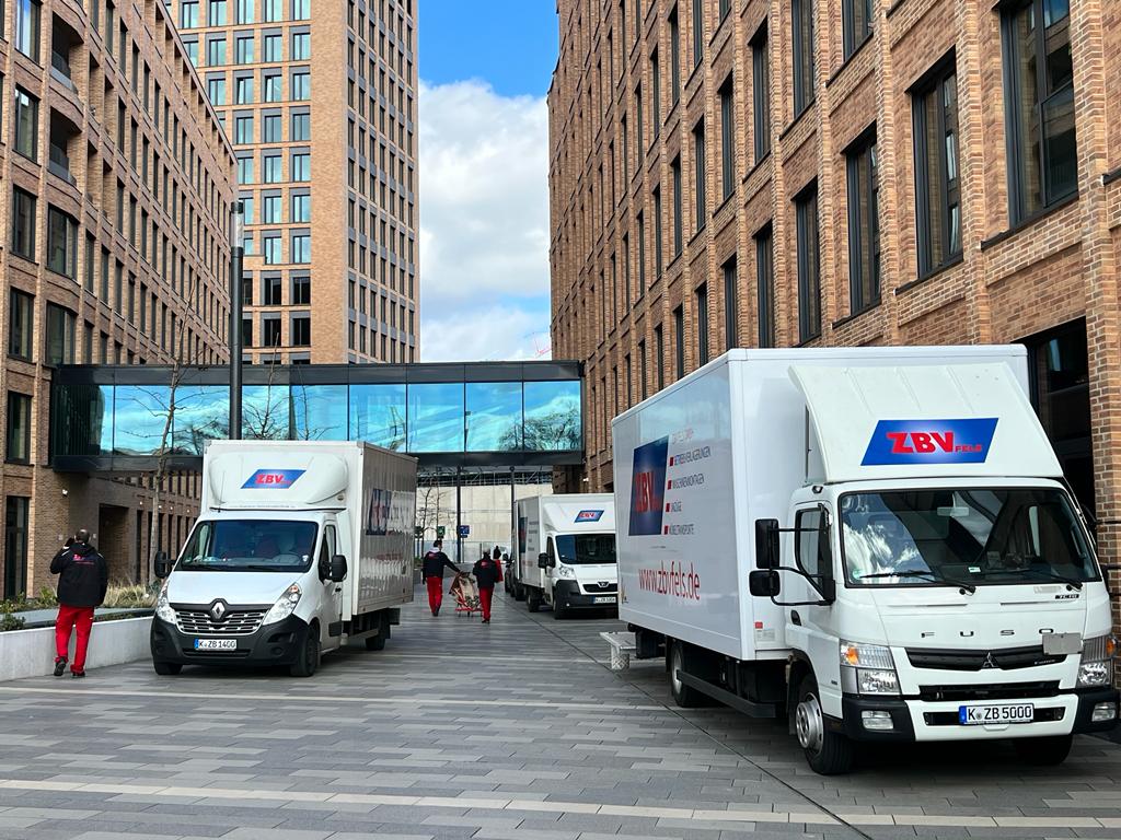 Zwei Lieferwagen in einer urbanen Straße zwischen hohen Backsteingebäuden mit Fußgängern.