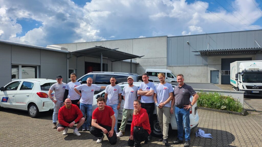 A group photo of eleven people in front of a company building with several vehicles in sunny weather.