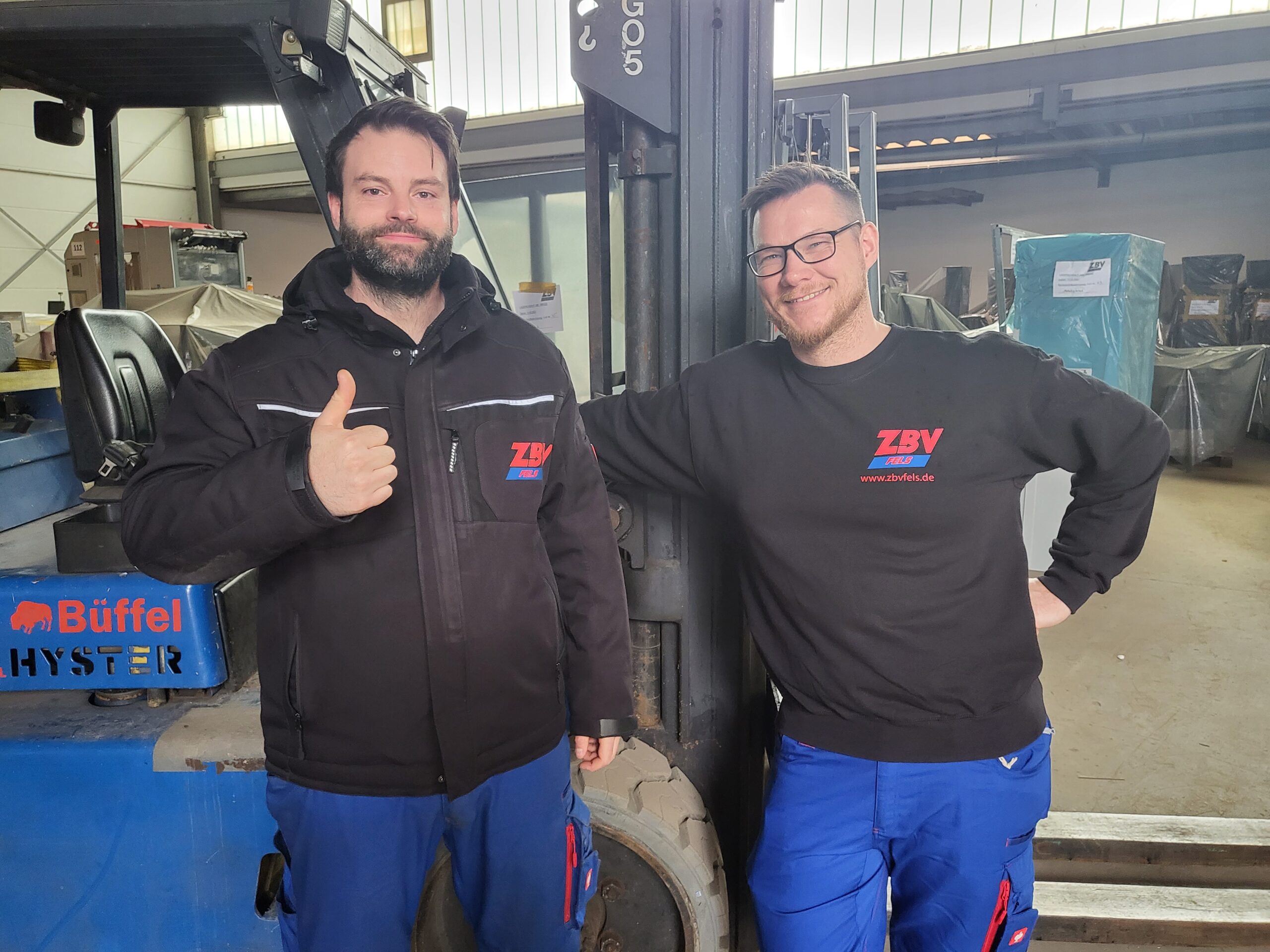 Zwei Männer in Arbeitskleidung in einer Werkstatt, einer zeigt Daumen hoch.