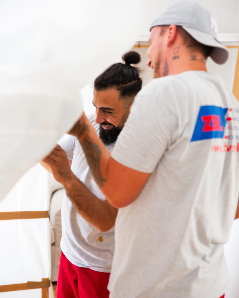 Two men working together on a piece of white fabric—one with a topknot and a beard, the other with a gray cap and tattoos.