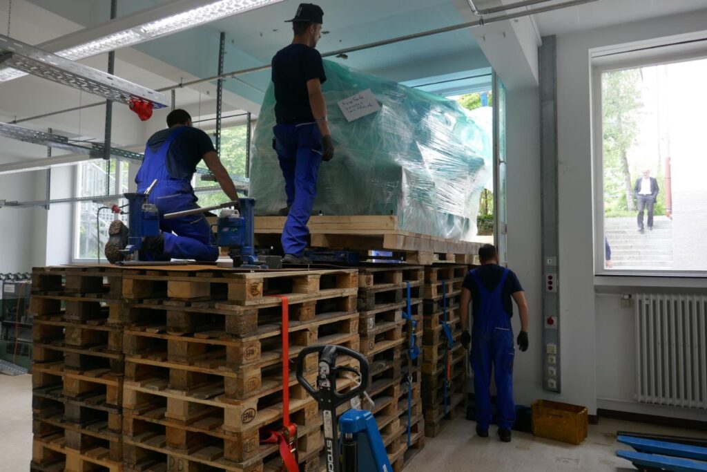 Drei Männer transportieren eine große Maschine durch ein Fenster, gestützt auf einen Turm aus Holzpaletten.