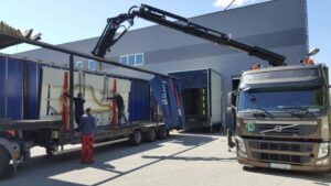 A truck with a crane is loading large glass panels into a warehouse; two workers are securing the load.