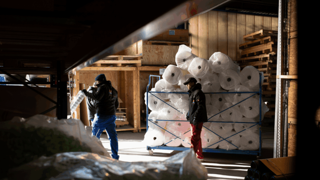 Two warehouse workers in work clothes are moving a cart with large rollers loaded with packaging materials through a warehouse—a light-filled industrial setting with shelves and pallets in the background.