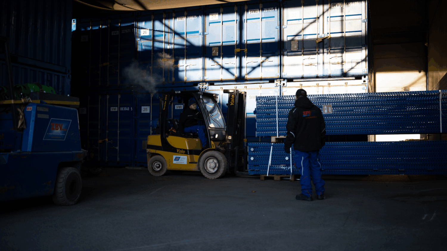 Gabelstaplerfahrer lädt schwere Metallregale vor Containern, während ein weiterer Mitarbeiter die Ladung koordiniert – professioneller Logistik- oder Montageservice in einer Lagerhalle.
