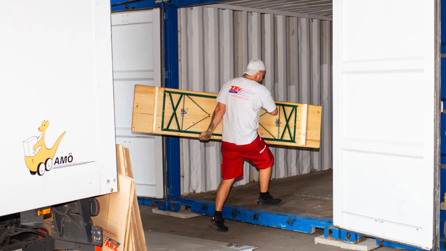„Mitarbeiter von ZBV Fels trägt ein zusammengeklapptes Holzmöbelstück in einen Selfstorage-Container – fachgerechte Einlagerung von Mobiliar für Umzug oder Zwischenlagerung.