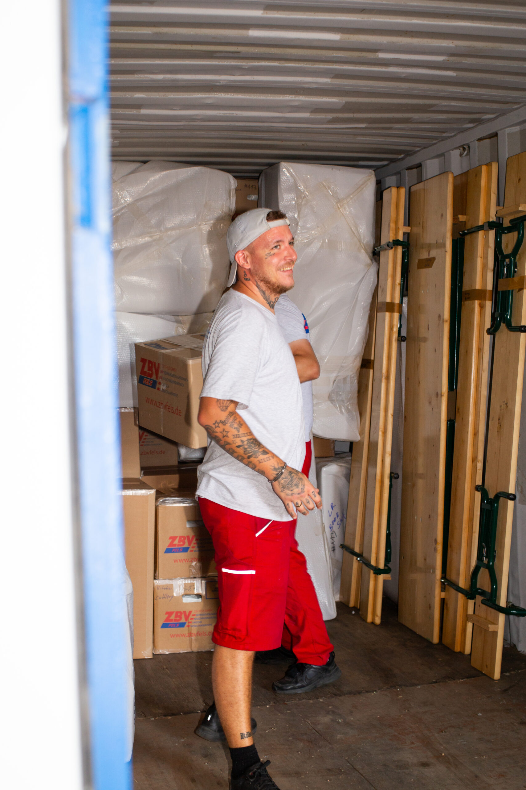 A man wearing red shorts and a white T-shirt is standing in a storage room filled with cardboard boxes and packing materials.