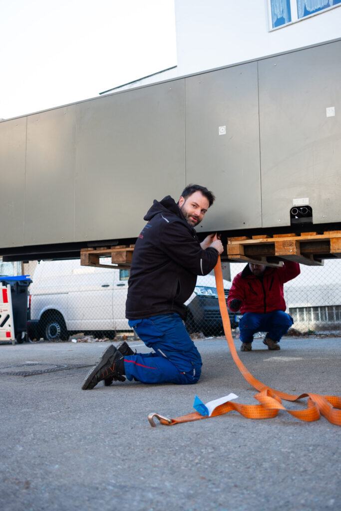 Two men are attaching an orange lifting strap to a large gray container on pallets.