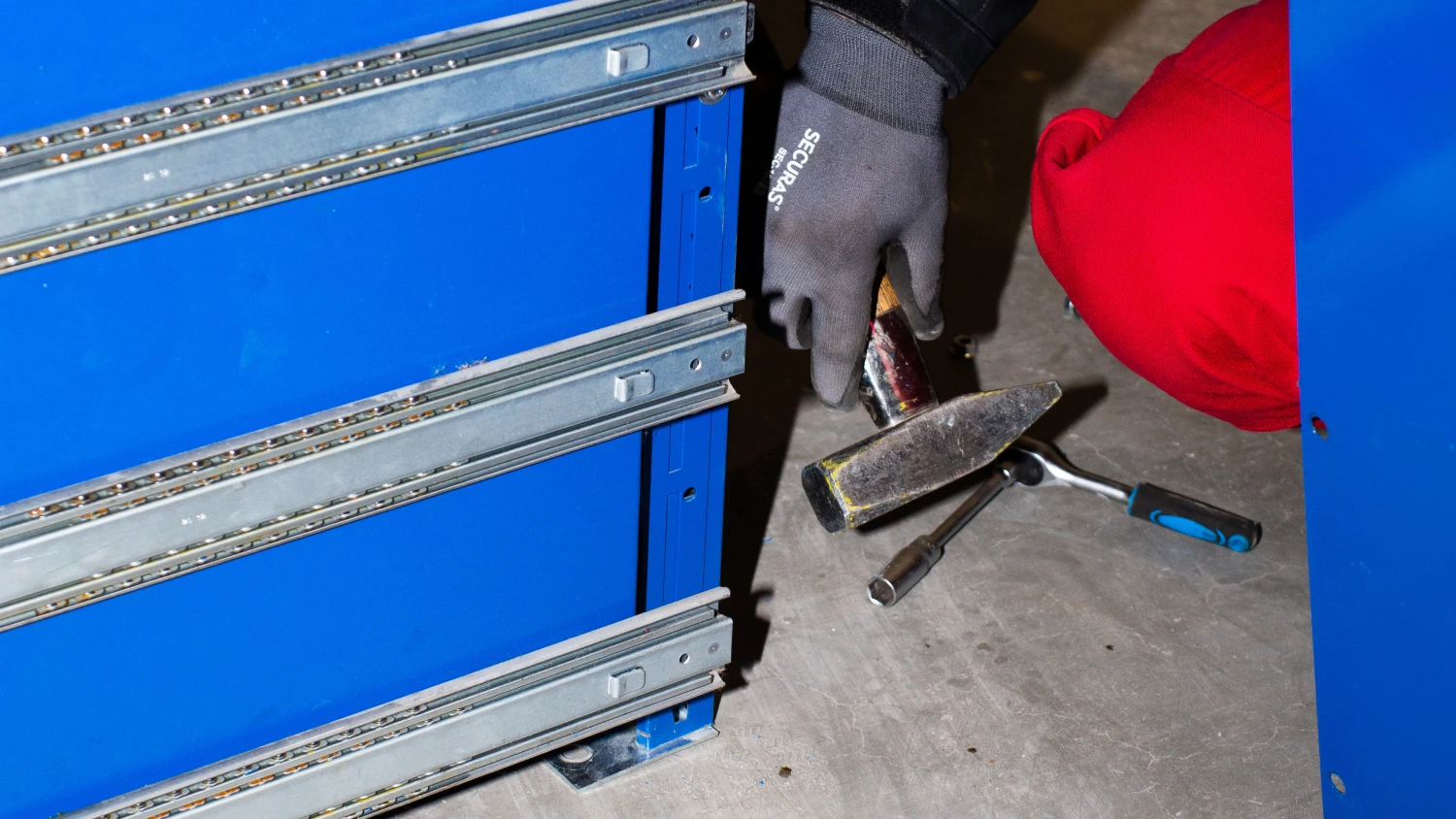 Detailaufnahme der Montagearbeit an einem blauen Regalsystem mit Werkzeugen und Arbeitshandschuhen