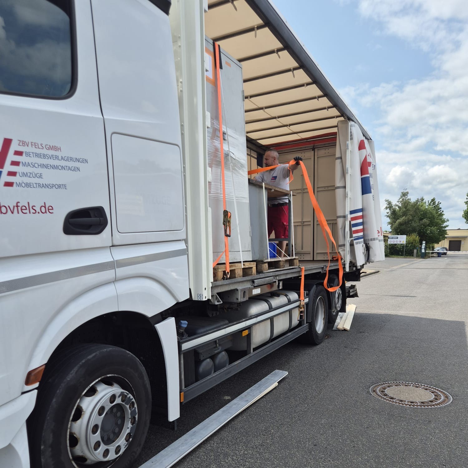 LKW mit geöffneter Ladefläche, beladen mit Paletten und gesichert mit orangenen Spanngurten.