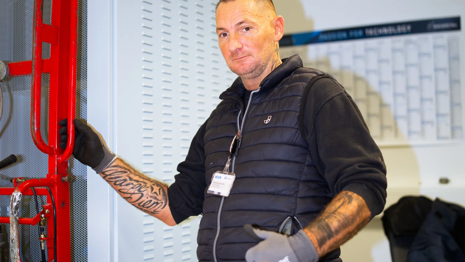 Mann mit Tattoos und Handschuhen in Arbeitskleidung vor rotem Rohr in Werkstatt.