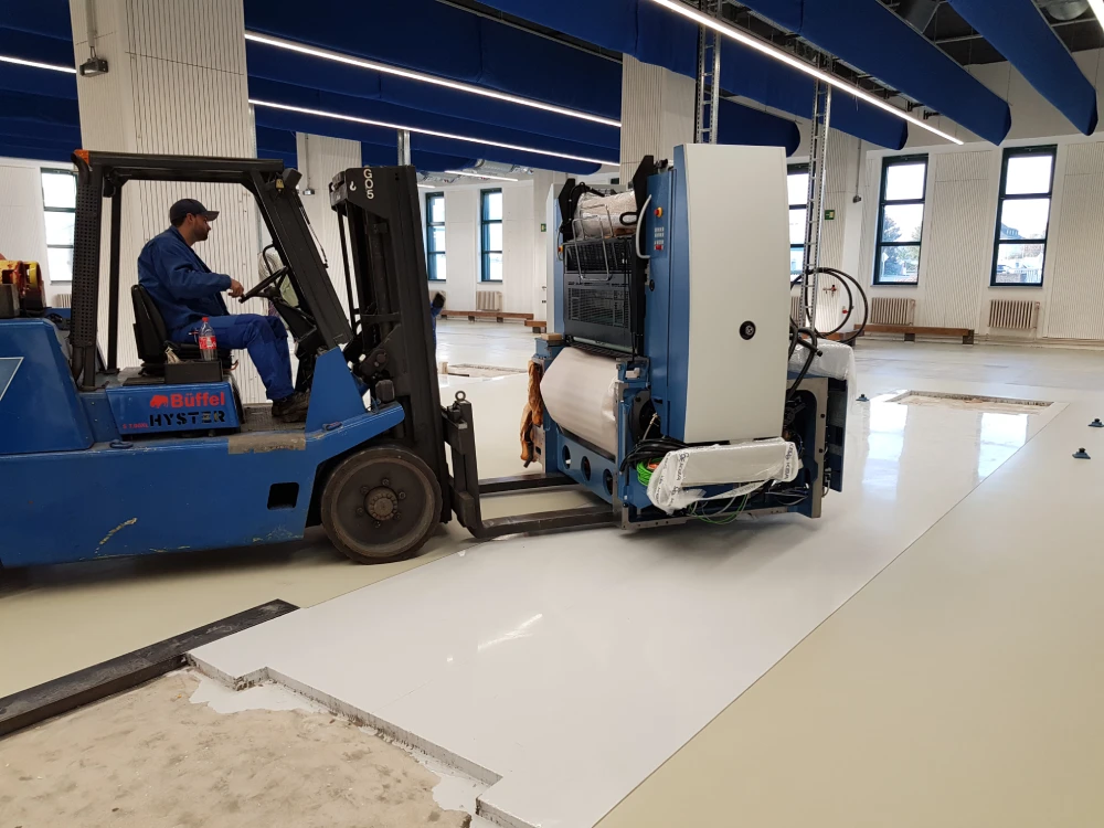 Gabelstapler transportiert große industrielle Maschine in einer Halle mit glänzendem Boden.