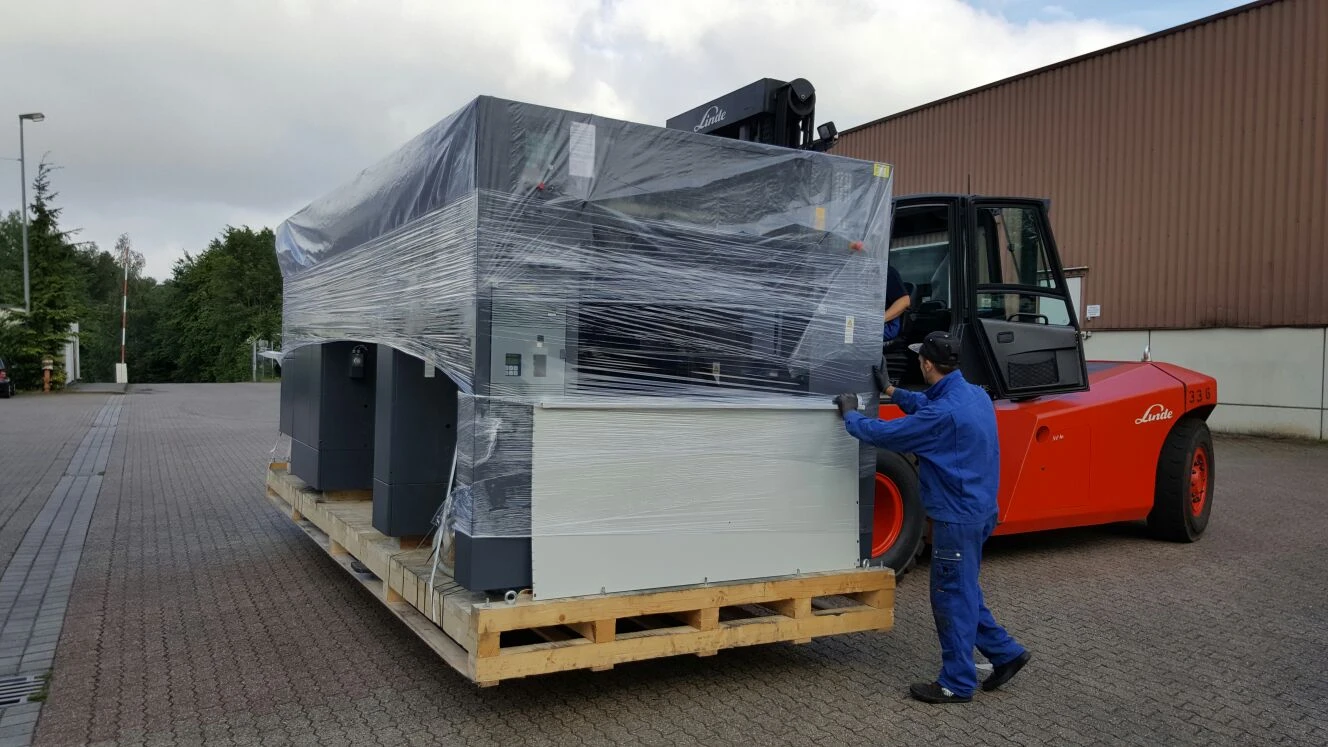 Gabelstapler lädt verpackte Maschine auf Palette, Arbeiter steht daneben.