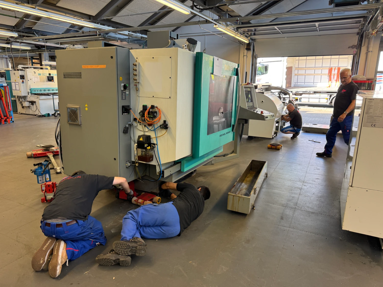 Zwei Männer reparieren eine große Maschine in einer Werkstatt, während zwei weitere Personen im Hintergrund arbeiten.