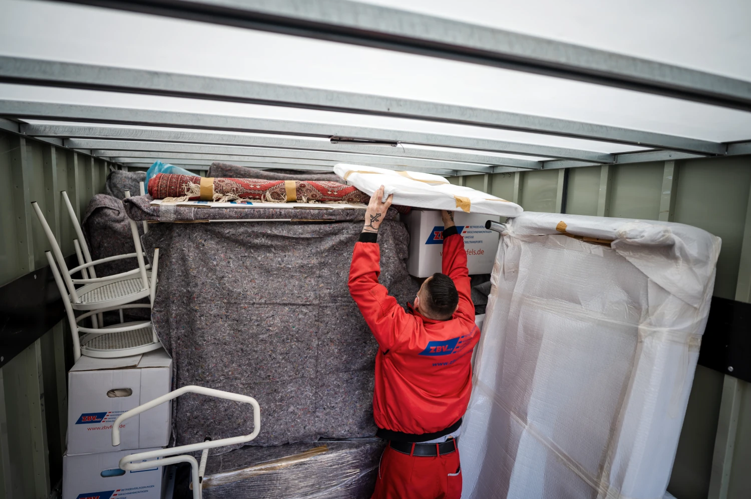 Person in roter Arbeitskleidung verstaut Möbel und Haushaltsgegenstände in einem Umzugscontainer.