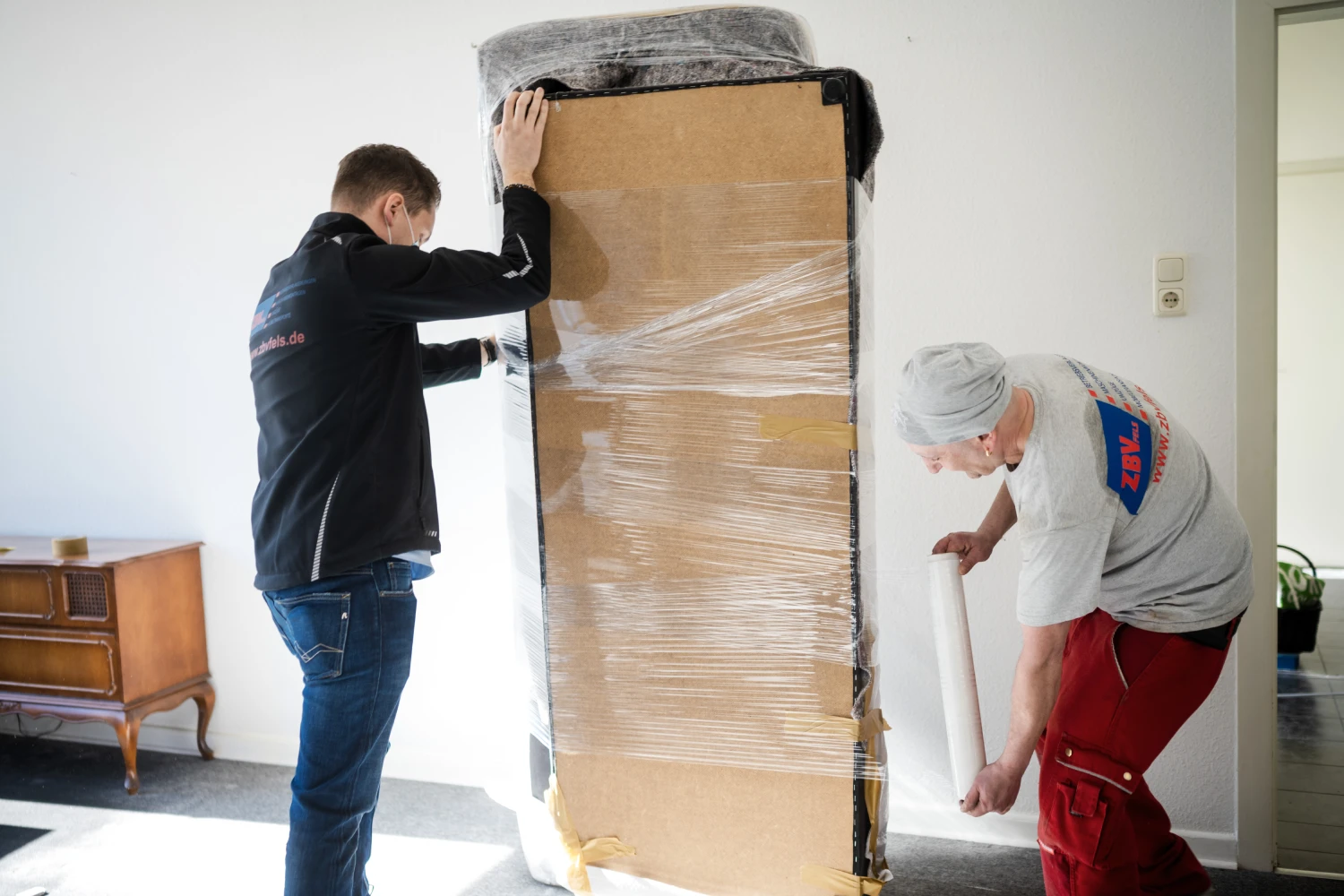 Zwei Männer verpacken einen großen Gegenstand mit Karton und Folie in einem Raum.