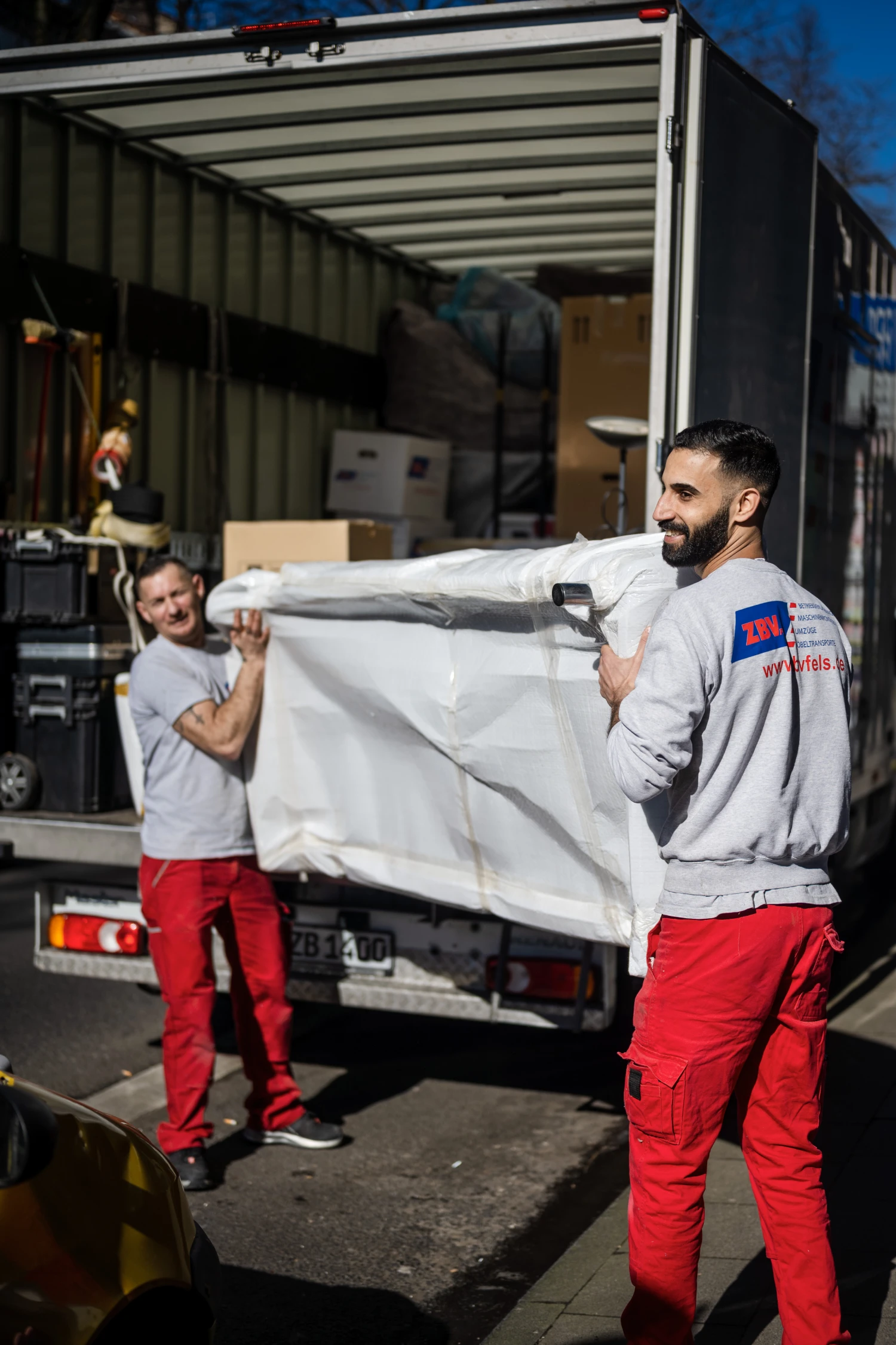 Zwei Männer in Arbeitskleidung tragen eine große weiße Verpackung aus einem LKW.