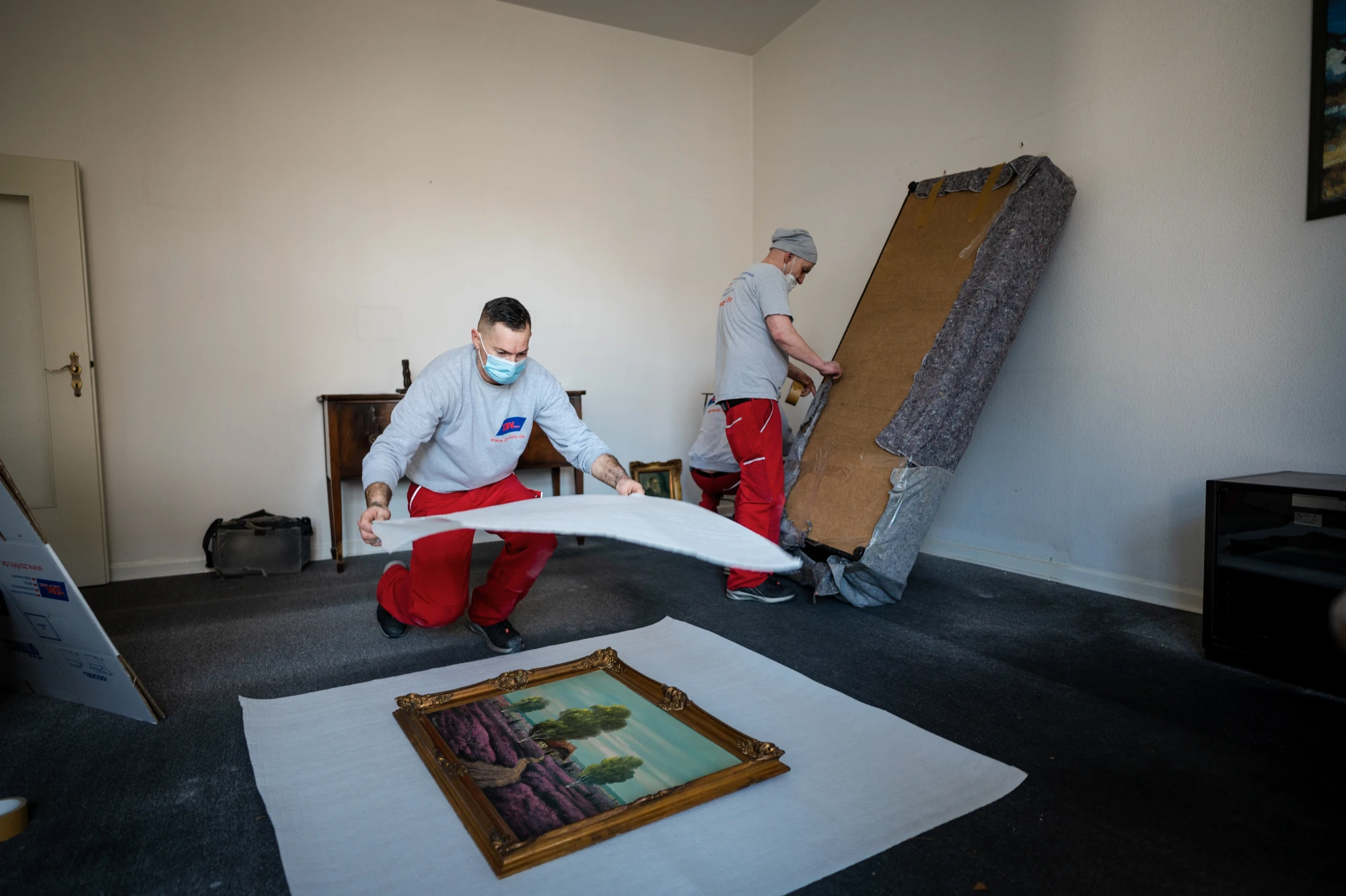 Zwei Männer mit Masken und roter Arbeitskleidung verpacken ein Gemälde in einem Raum.