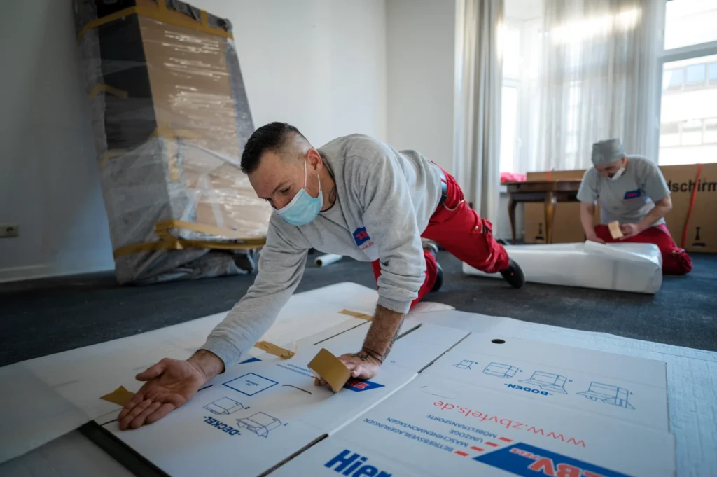 Zwei Männer mit Mundschutz verlegen Bodenbeläge in einem Raum.