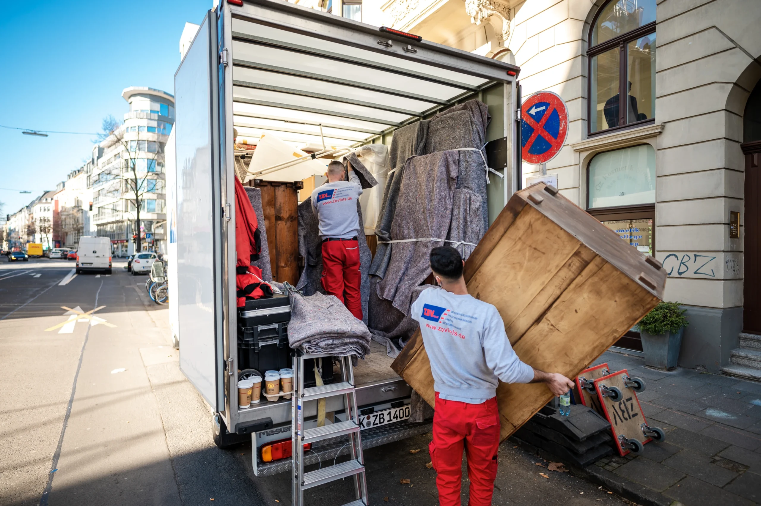 Zwei Umzugshelfer laden Möbel aus einem LKW vor einem Wohnhaus in einer Stadtstraße.