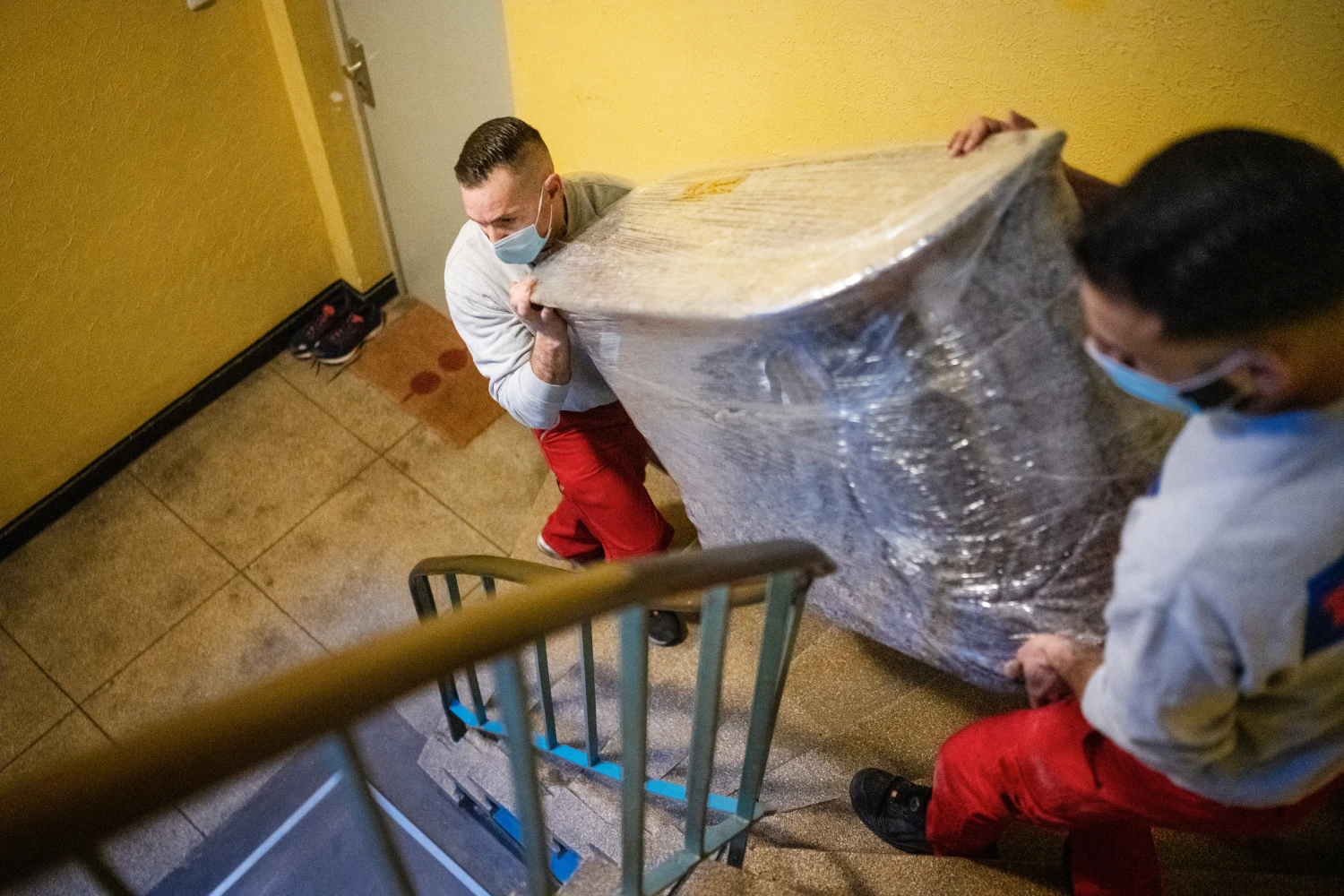 Zwei Männer mit Masken tragen einen großen, in Folie eingewickelten Gegenstand eine Treppe hinauf.