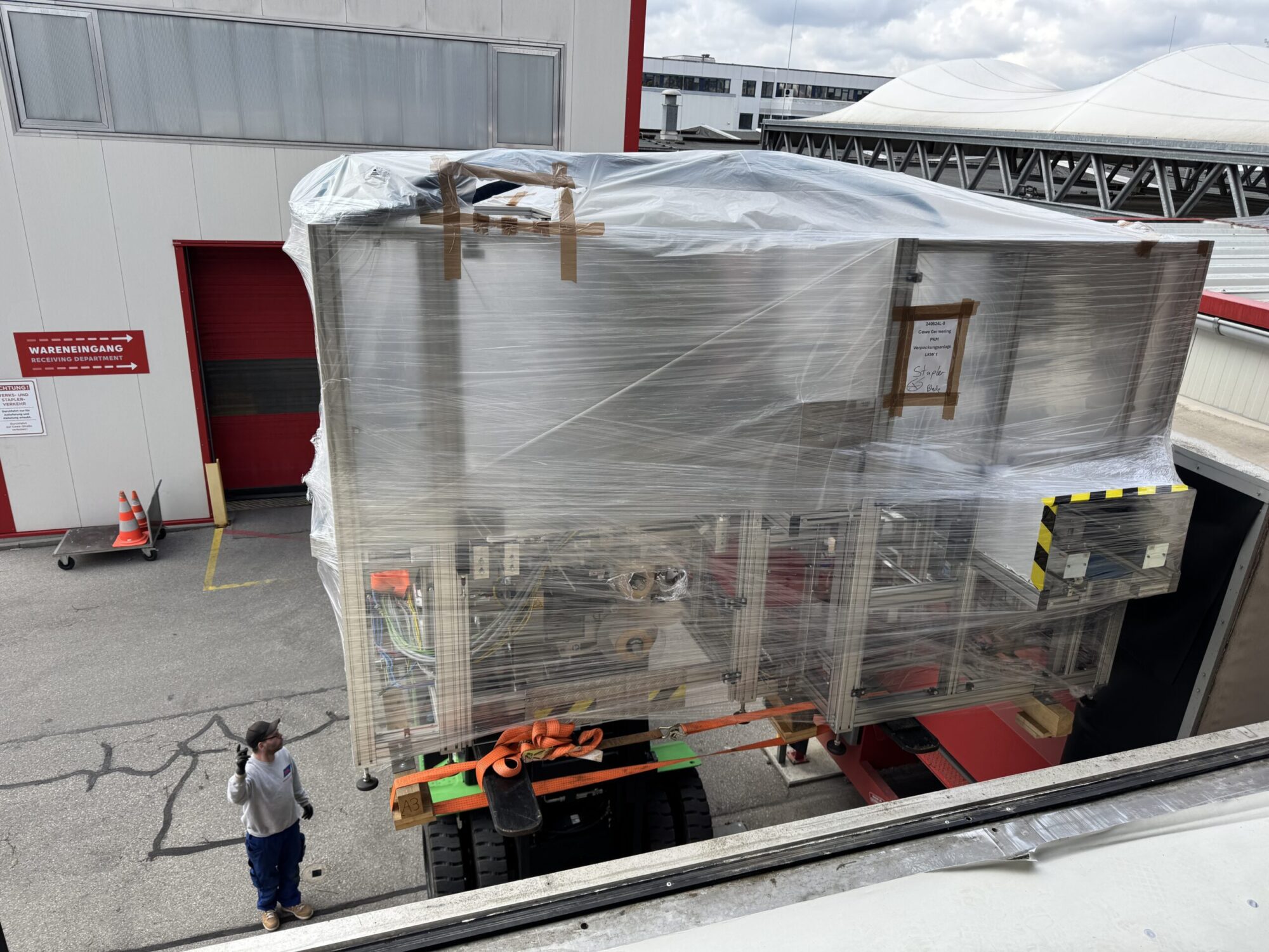 Großes, in Folie verpacktes Objekt auf einem LKW-Anhänger vor einem Gebäude, zwei Personen daneben.