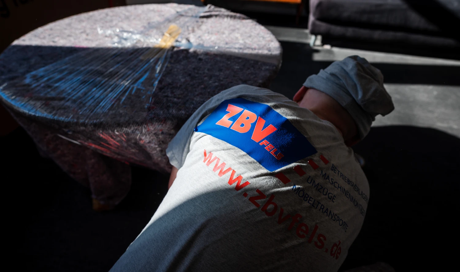 Person in Arbeitskleidung mit ZBV-Logo, die auf dem Boden arbeitet, neben einem runden Tisch mit Folie bedeckt.