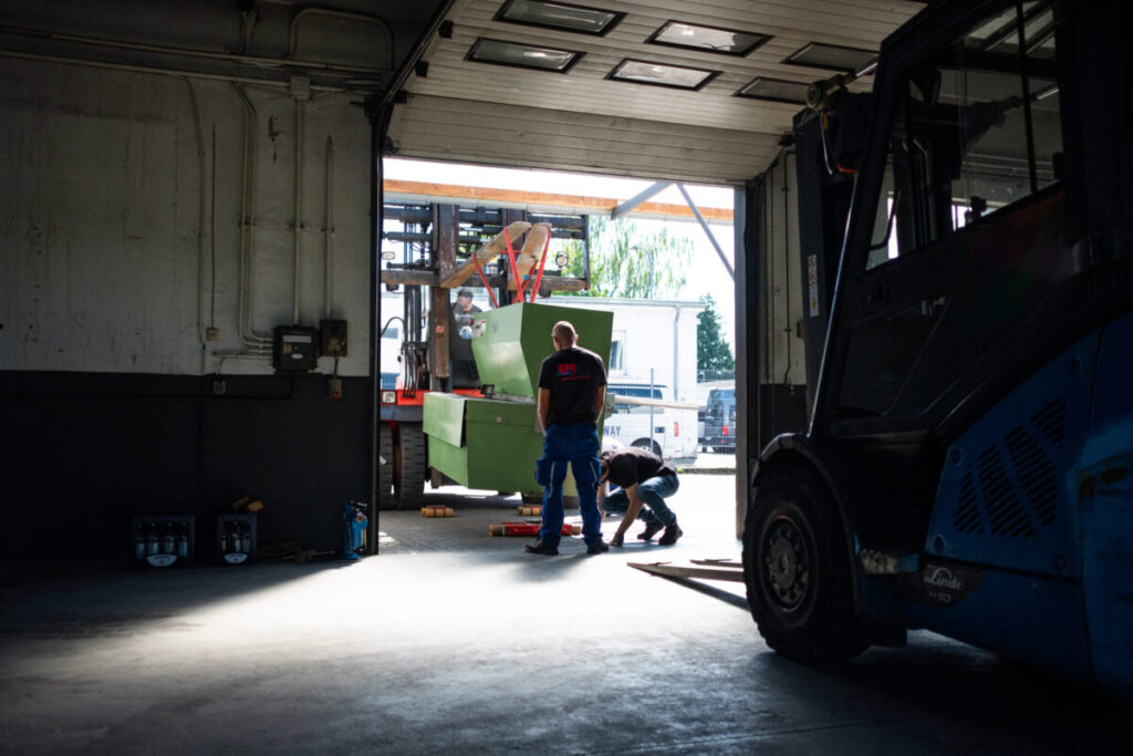Workers secure a large green machine as it is being transported by forklift to the warehouse exit