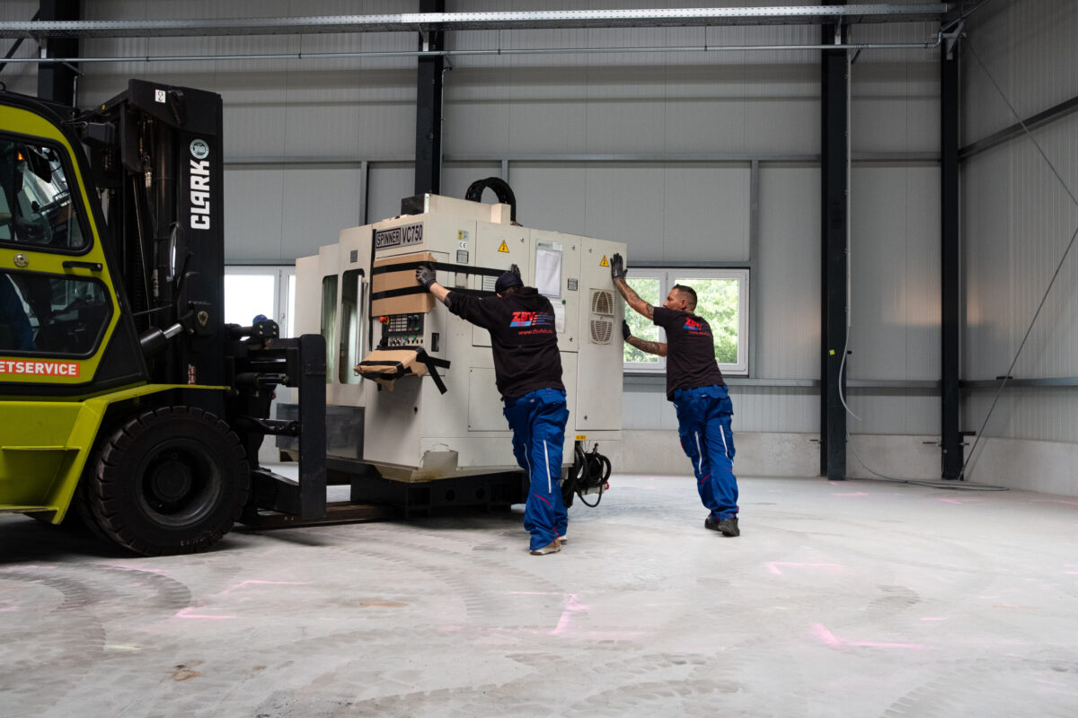 Zwei Mitarbeiter positionieren Maschine mit Gabelstapler in Werkhalle