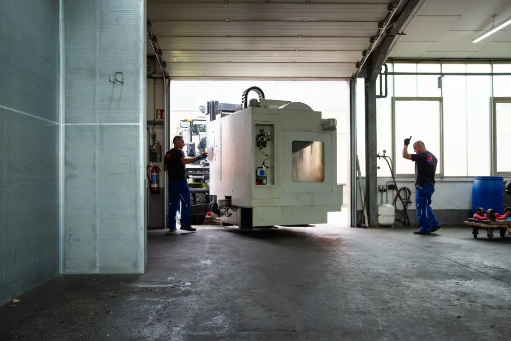 Zwei Arbeiter bewegen eine große Maschine in einer Werkstatt mit offener Garagentür.