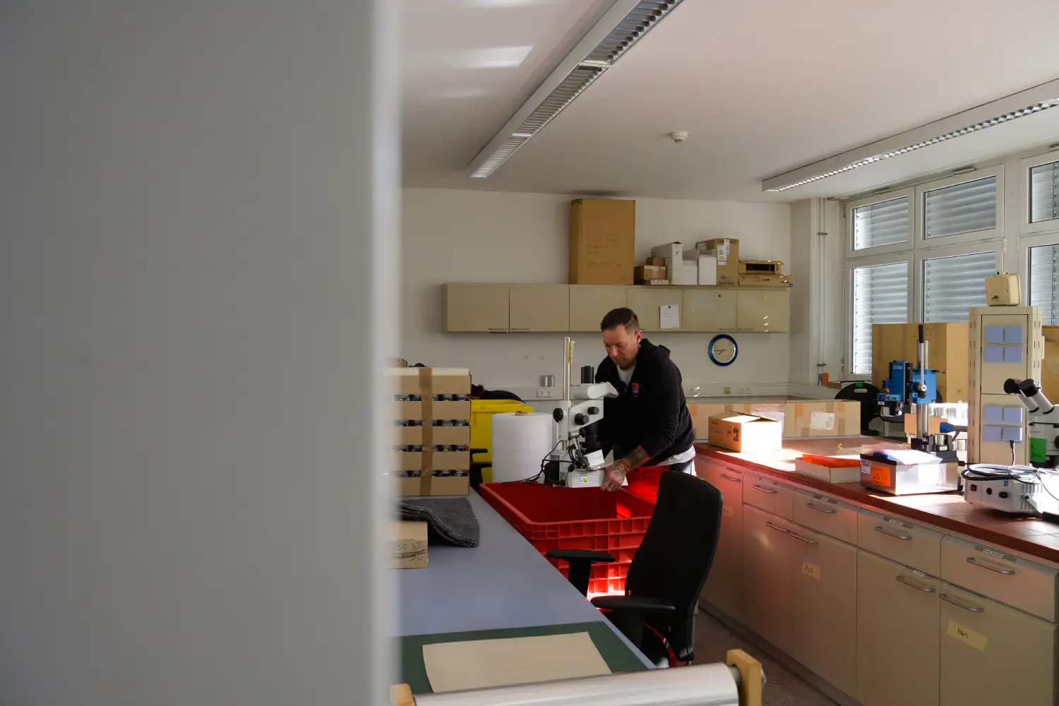 Person arbeitet mit Mikroskop in einem Labor mit Schränken und Regalen.