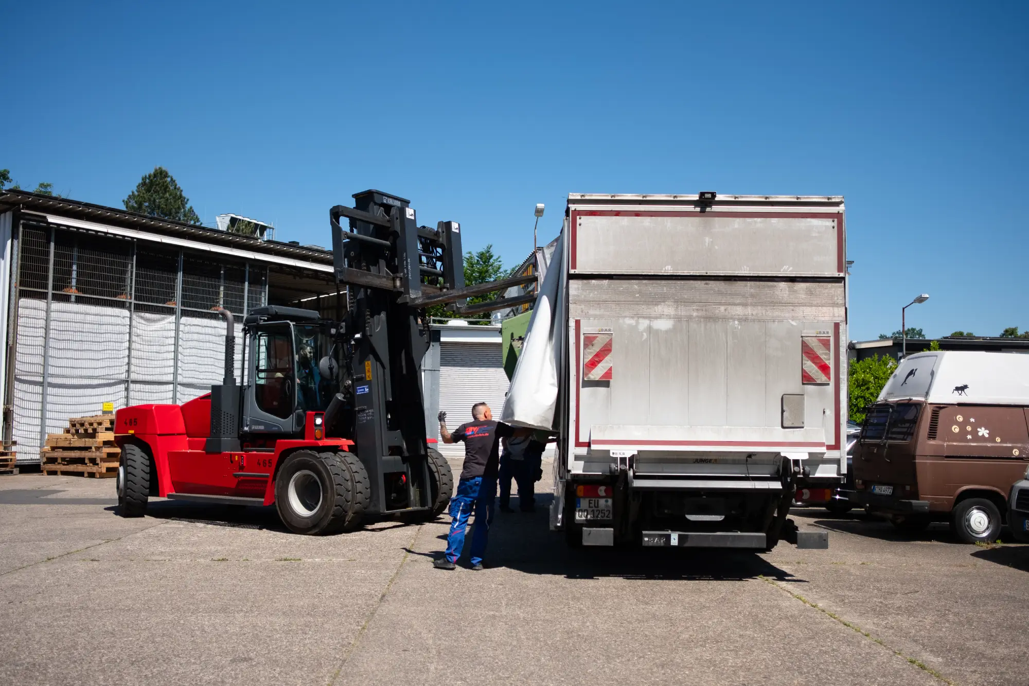 Gabelstapler lädt eine große weiße Tonne auf einen LKW, zwei Männer helfen dabei.