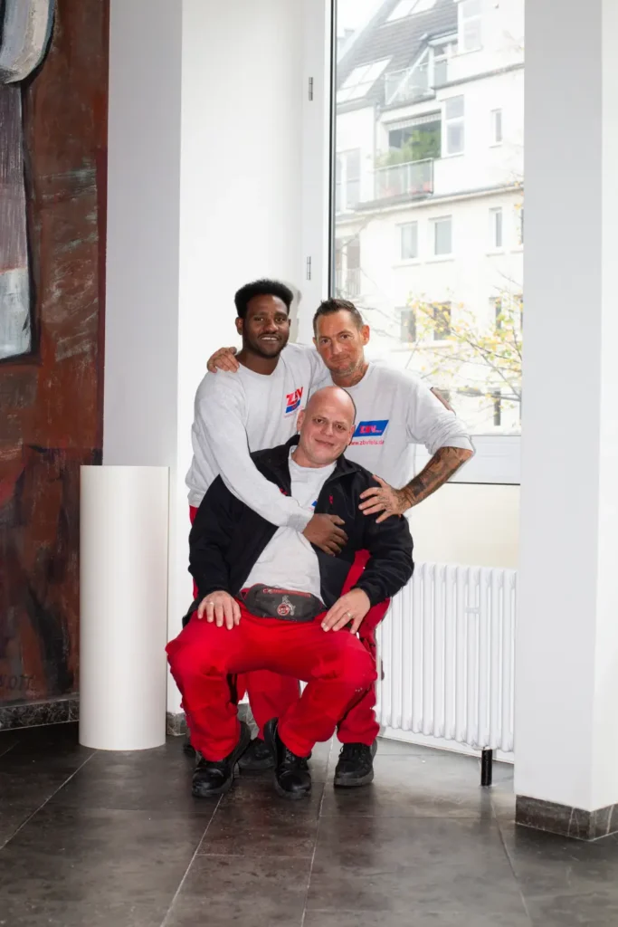 Three men in a brightly lit room; two are standing behind a man who is sitting, and they are all smiling.